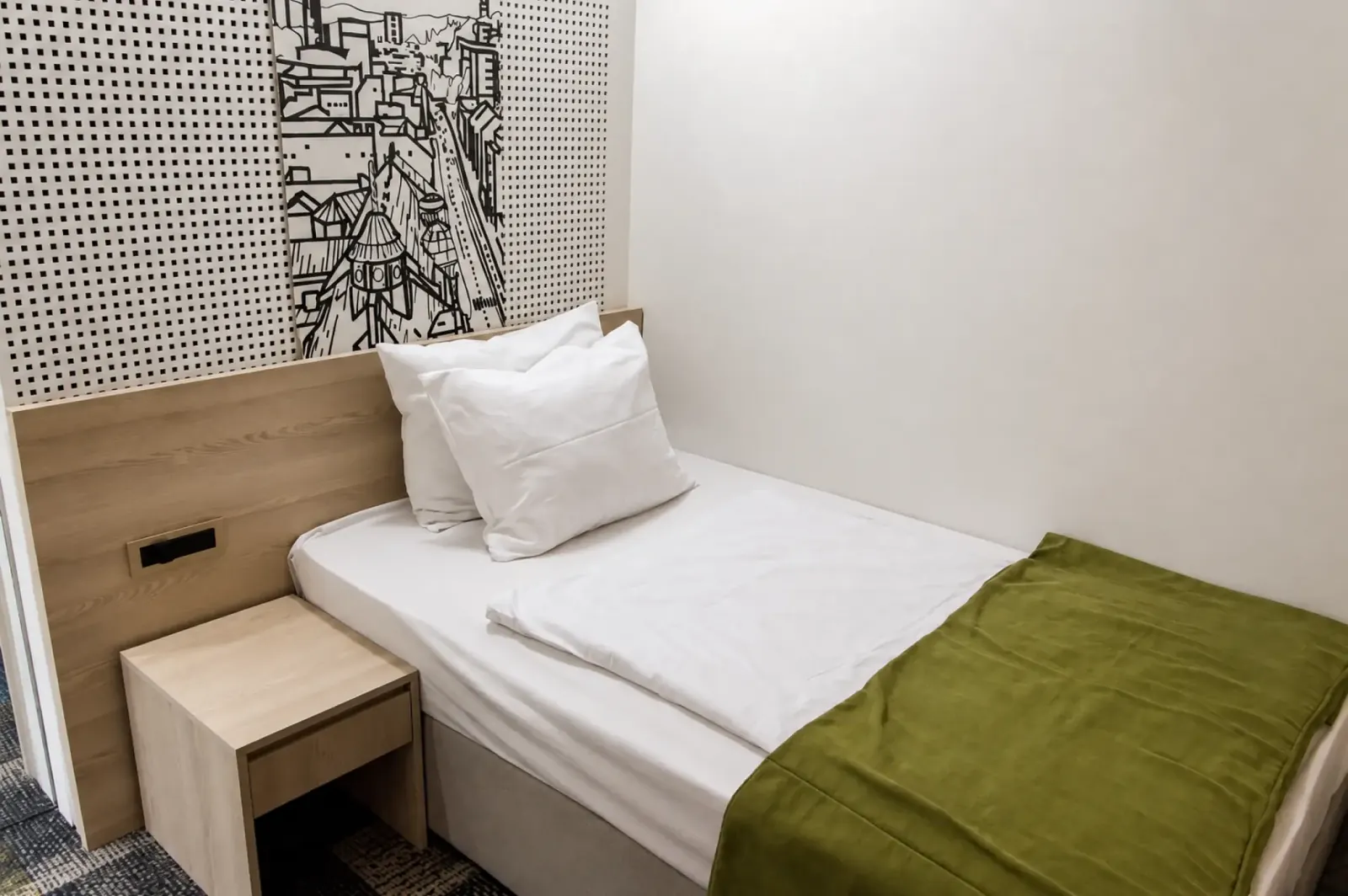 Single bed with white pillows, green cover and city mural at Hotel Cosmopolit